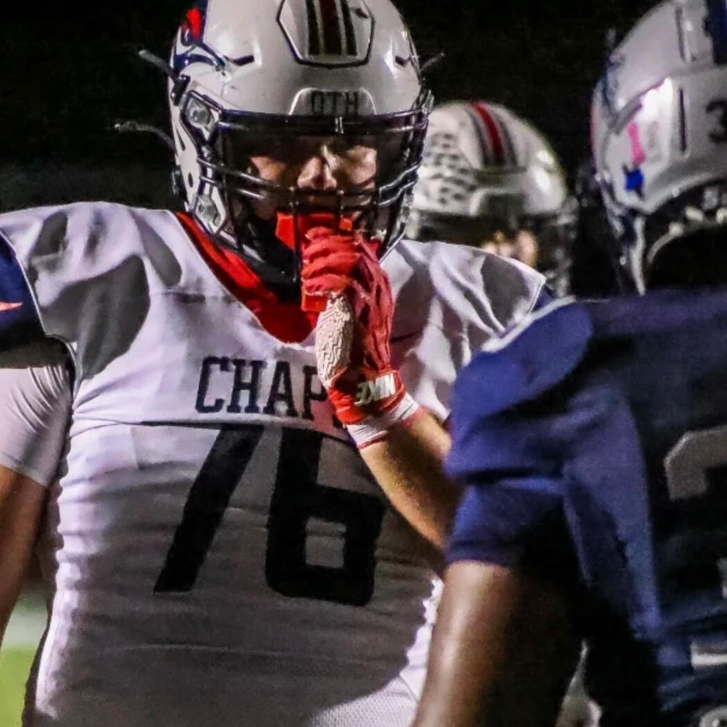 Aubrey High School football player Charles Mulkey (#76), Class of 2028, stands at the line of scrimmage during a nighttime game wearing a white Chaparrals uniform and helmet, gripping his mouthguard while facing an opposing player before the snap.